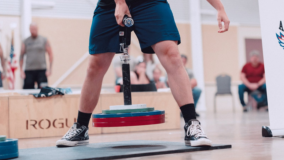 Lifter at a grip competition pulling a Country Crush Raptor rolling handle loaded with plates on a loading pin and swivel carabiner, spectators watching in the background.